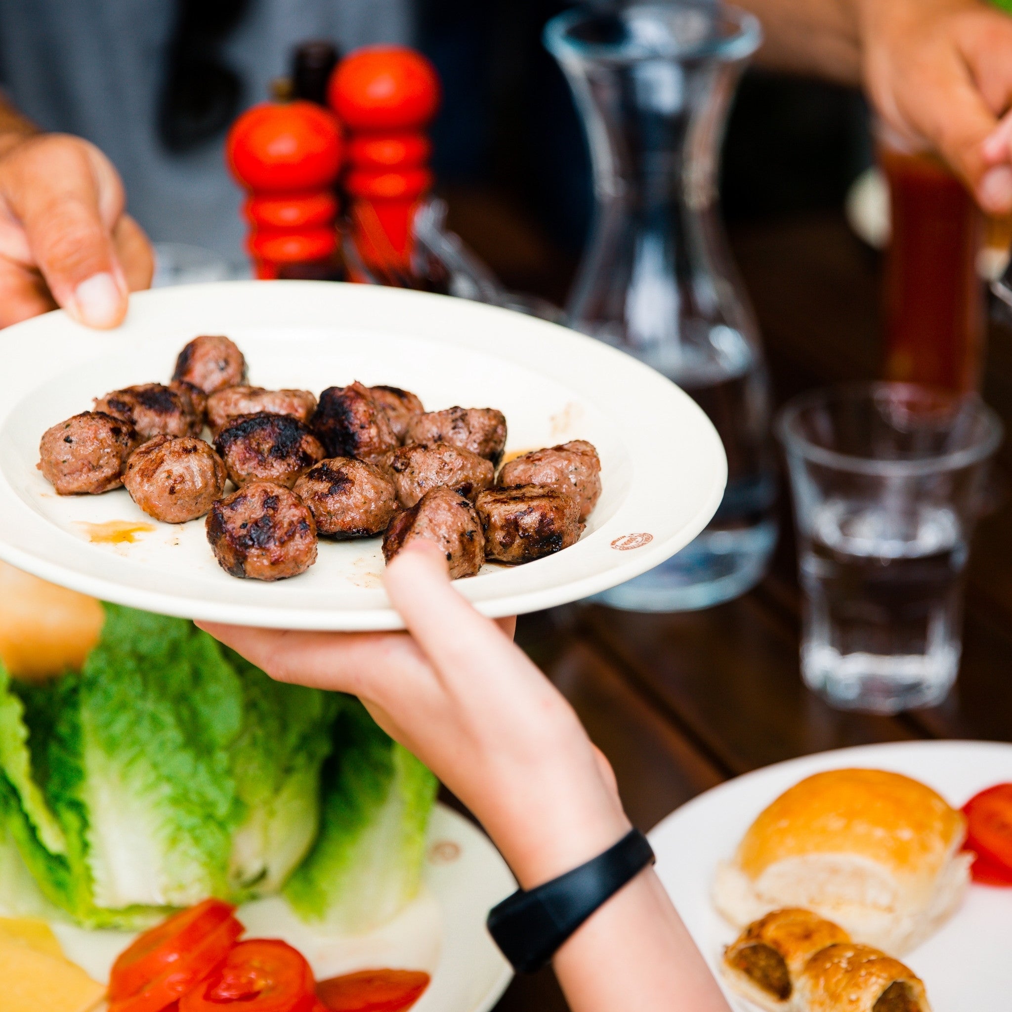 Person holding a plate of grilled meatballs with a blurred background of a dining table.