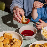 Two people eating food with a bowl of red sauce on a table.