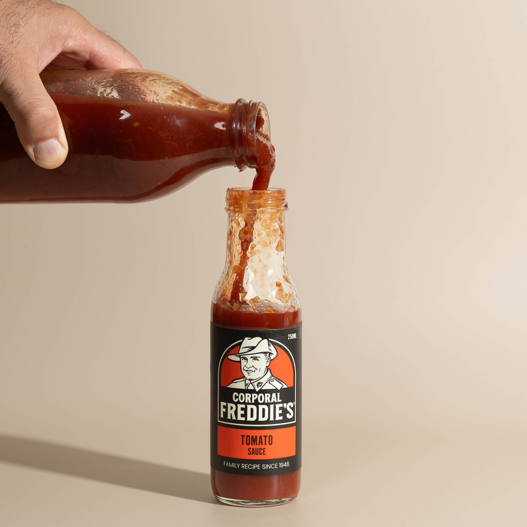 Hand pouring tomato sauce into Corporal Freddie's Tomato Sauce on a beige background.