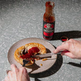 Person cutting into a pastry with red sauce on a plate with a bottle of 'Corporal Freddie's' hot sauce.