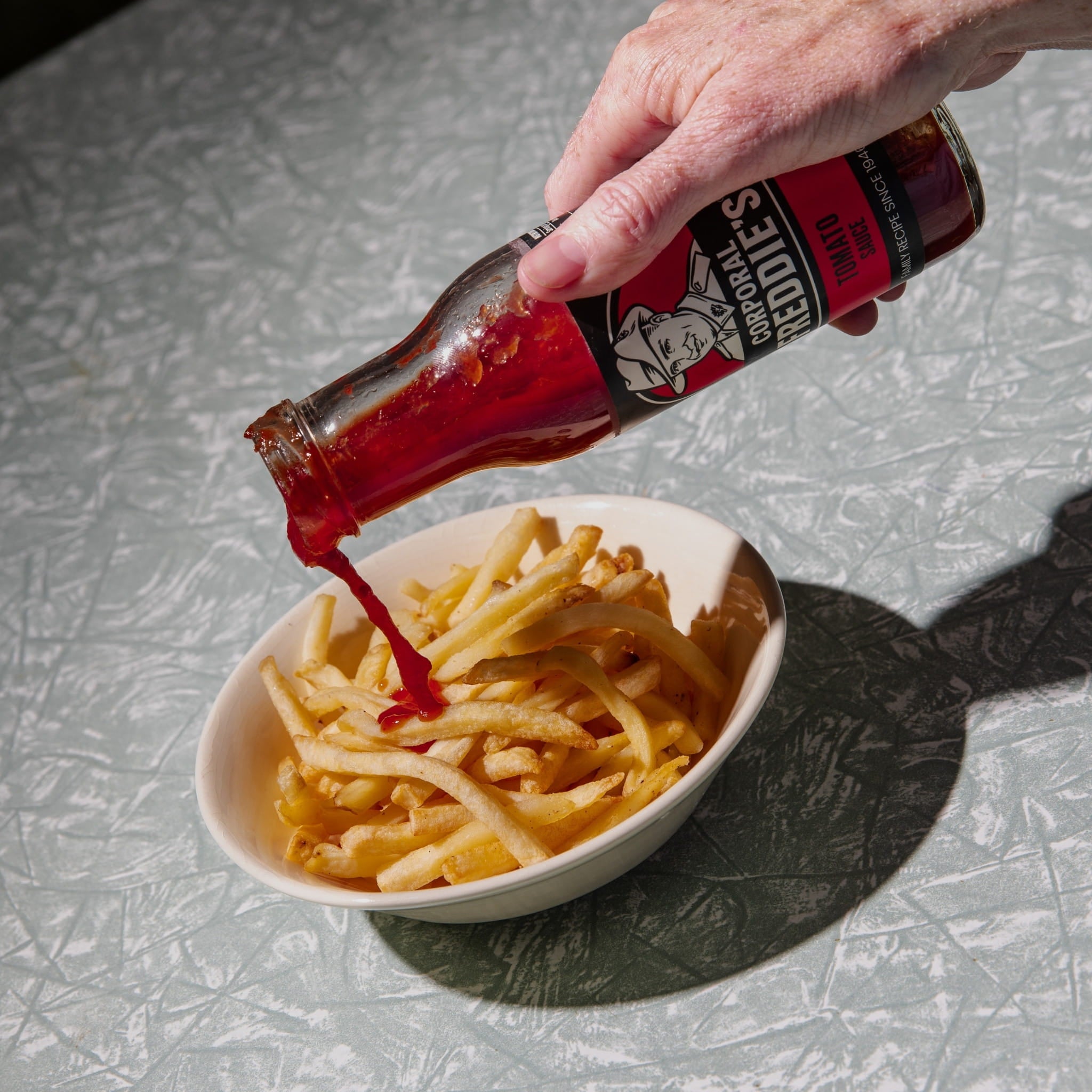 Hand pouring Freddie's Tomato sauce onto a bowl of fries