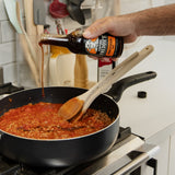 Person pouring Corporal Freddie's Worcestershire sauce from a bottle into a pan with a wooden spoon on a stove.