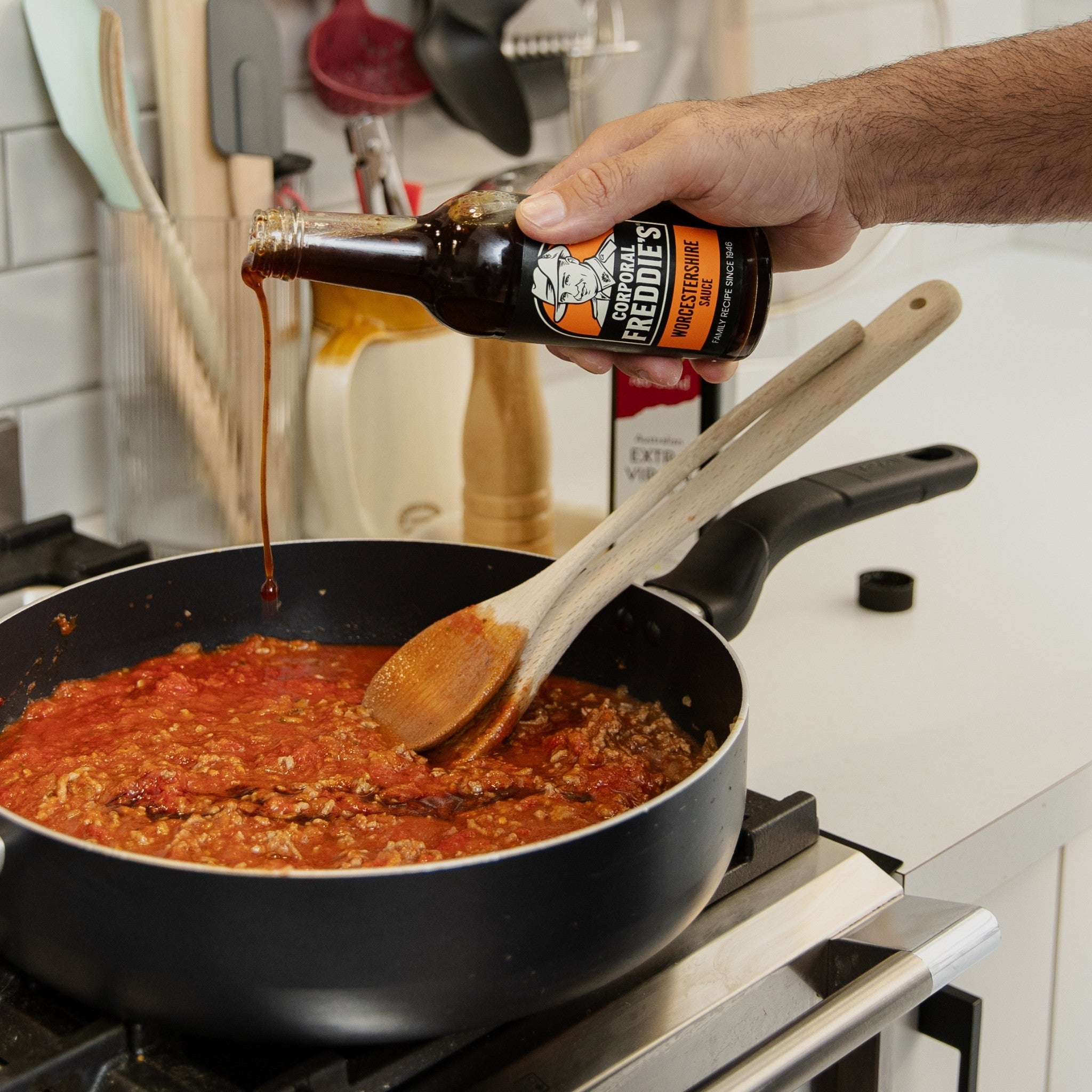 Person pouring Corporal Freddie's Worcestershire sauce from a bottle into a pan with a wooden spoon on a stove.