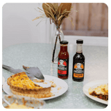 Two bottles of Colonel Freddie's hot sauce on a table with a pie and other food items.