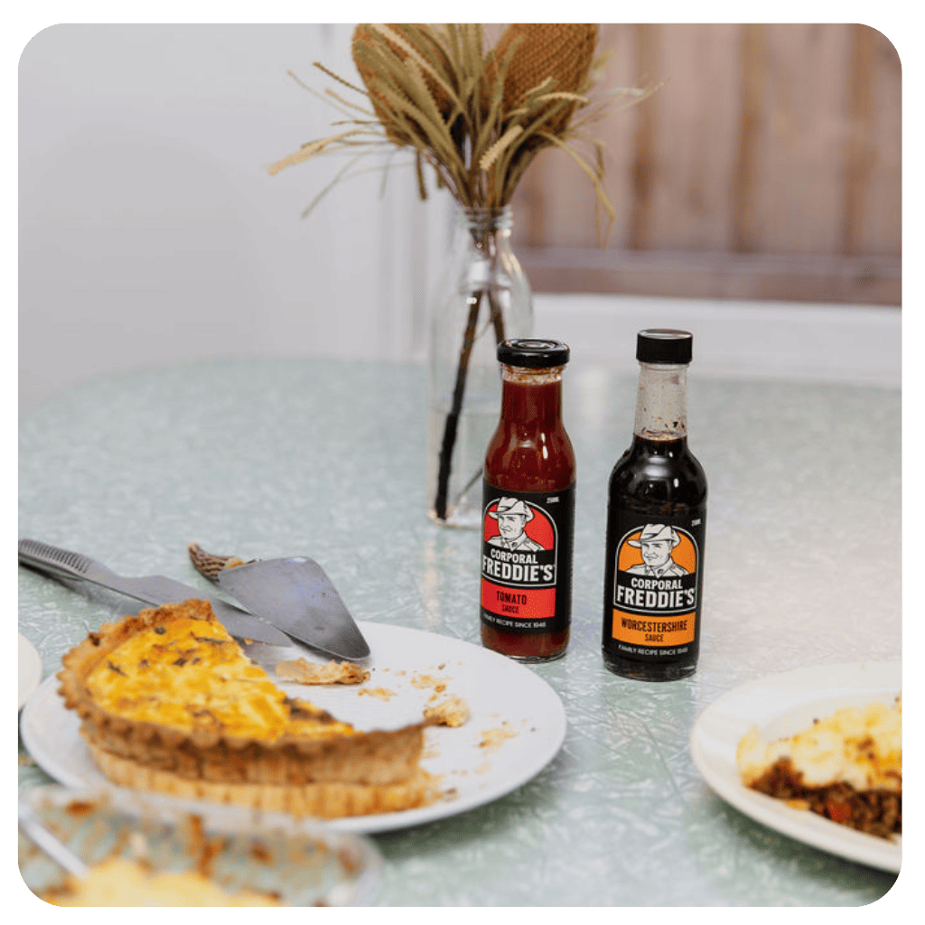 Two bottles of Colonel Freddie's hot sauce on a table with a pie and other food items.